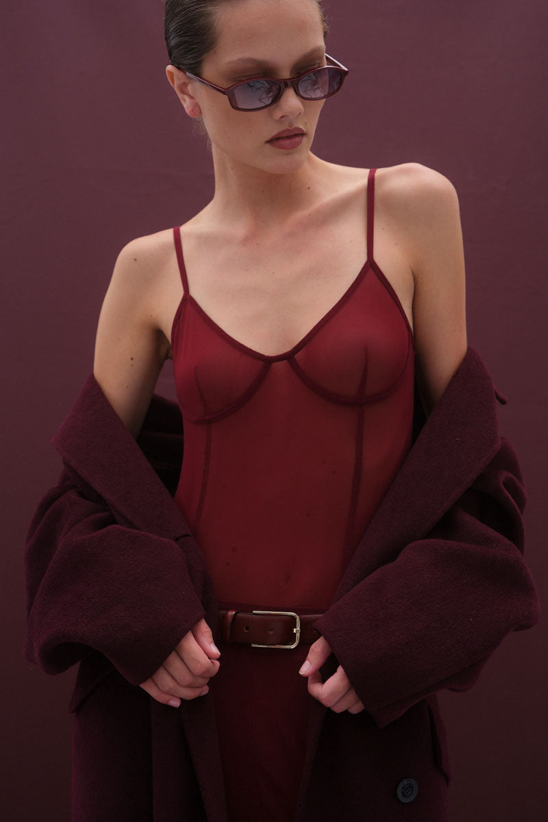 Woman wearing a burgundy bodysuit with thin straps and a matching coat against a purple background