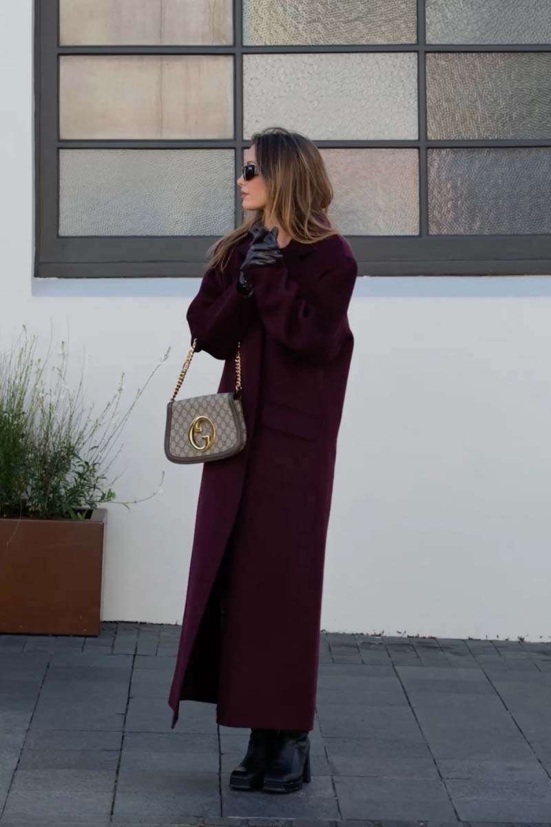 Woman in a long dark coat holding a Gucci handbag against a white wall with a window.
